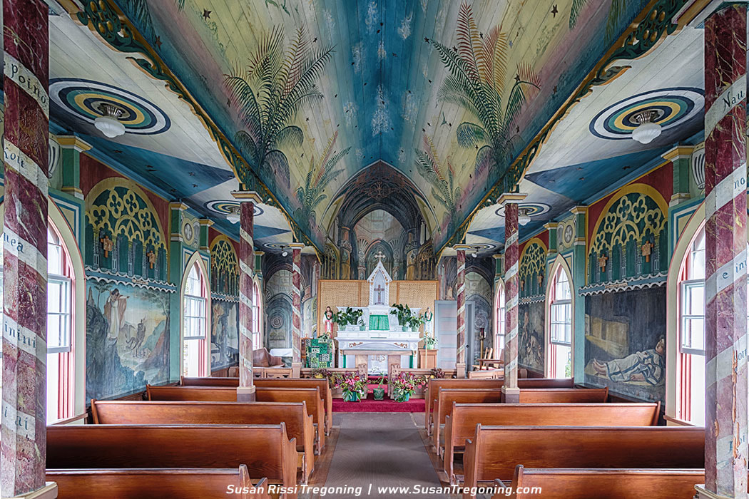 Interior of St. Benedict Painted Church with wooden pews facing a small altar decorated with green cloths and flowers. The ceiling is painted with a sky scene of blue tones, palm fronds, and stars, while the walls feature colorful religious murals and faux‑marble arches. Natural light enters through arched windows, illuminating the vivid artwork covering nearly every surface.
