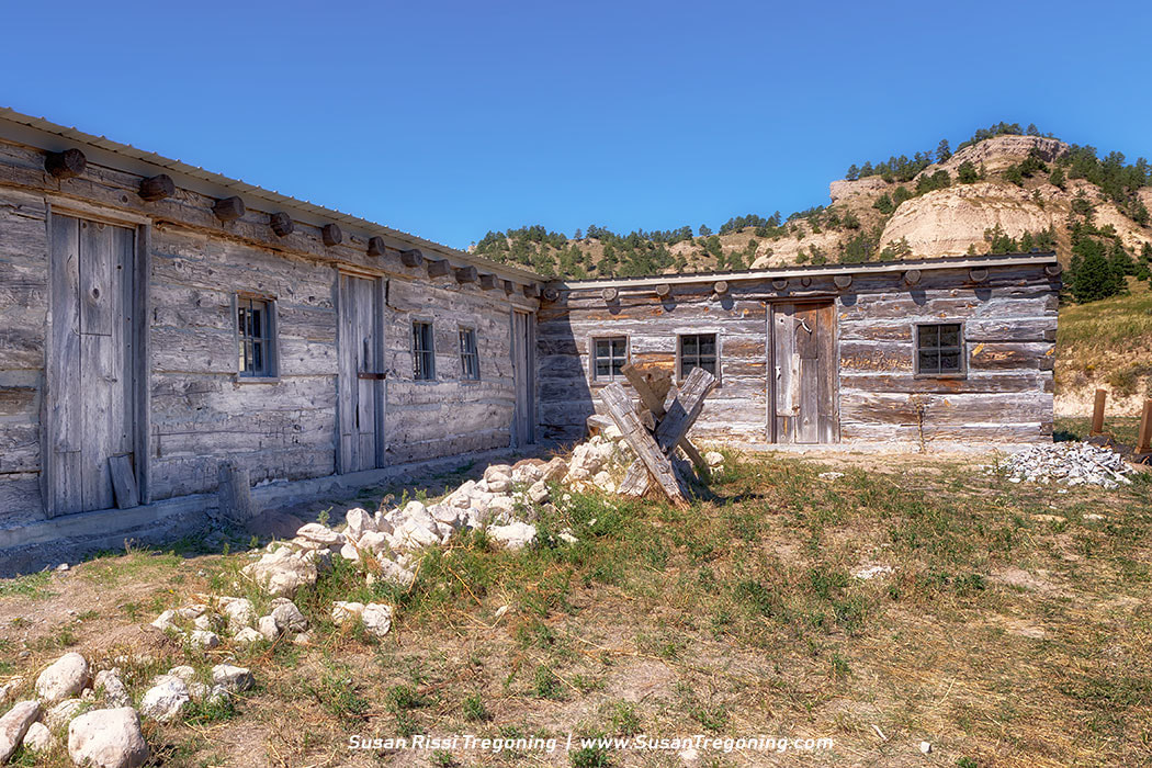 A recreated log trading post stands in a grassy clearing with hills and scattered trees in the background under a clear blue sky.
