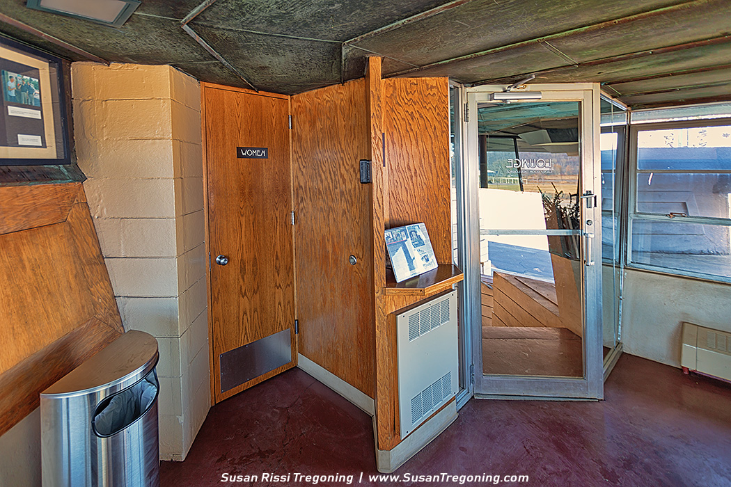 A small interior vestibule features a wooden door labeled “Women” beside another wooden door with a small display shelf holding a framed item. To the right, a glass door marked “Lounge” leads to another room. The space includes wood paneling, concrete block walls, a metal trash can, and a textured ceiling.