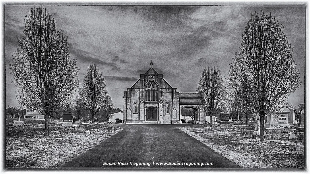 A black‑and‑white photograph of the historic Goddard Chapel in Marion, Illinois, centered at the end of a straight paved walkway in Rose Hill Cemetery. Leafless trees line both sides of the path, with rows of gravestones visible beneath them. Two matching headstones in the foreground read “GOODALL.” The Gothic‑style stone chapel rises in the distance with pointed arches and ornate window tracery, framed by a cloudy, dramatic sky.