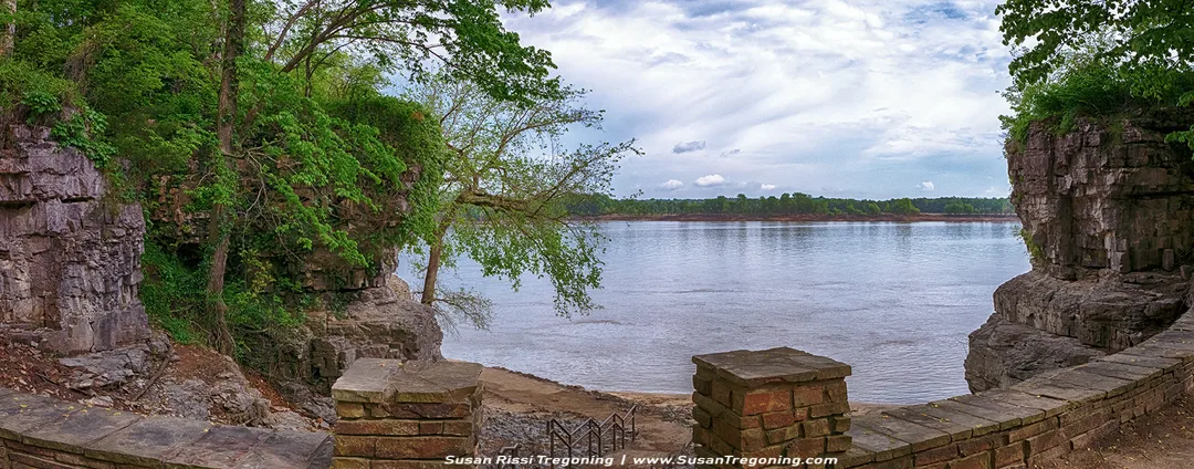 A panoramic view of the Ohio River is seen from a stone overlook at the top of a stairway leading down toward a cave. Rocky cliffs and dense green vegetation frame the river as it stretches into the distance beneath a partly cloudy sky.