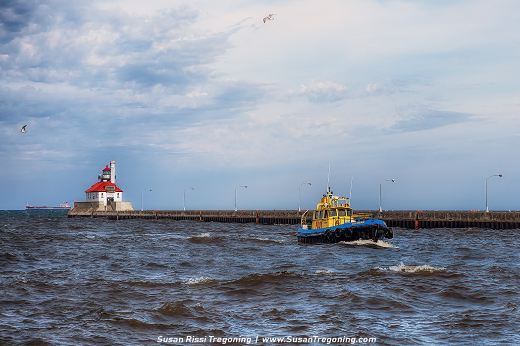The yellow and blue pilot tug Sea Bear moves through choppy water toward the Duluth Ship Canal, with the white lighthouse on the pier to the right and a large cargo ship visible on the horizon. Several lampposts line the pier, and seagulls fly above the waves under a partly cloudy sky.