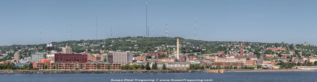 A panoramic view of Duluth, Minnesota, showing the city built along the Lake Superior waterfront with neighborhoods rising up the hillside. Numerous communication towers line the ridge above the city. The scene includes a mix of modern buildings, older brick structures, and industrial areas, with calm water in the foreground and a clear blue sky overhead.
