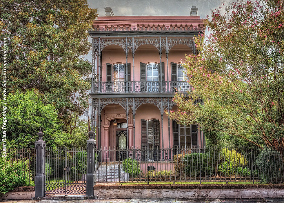 A two‑story historic house with a pink exterior features tall shuttered windows, ornate wrought‑iron balconies, and a central doorway framed by decorative ironwork. Lush greenery surrounds the home, and a black iron fence encloses the property, highlighting its symmetrical Victorian‑era design.