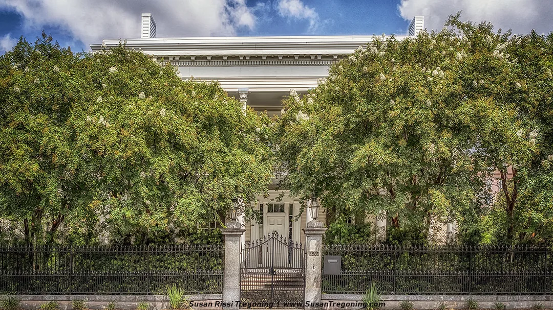   
A stately white building with a symmetrical façade features tall classical columns, a decorative cornice, and an entrance framed by stone pillars topped with lanterns. Lush green trees partially obscure the structure, and a black wrought‑iron fence and gate enclose the front of the property.