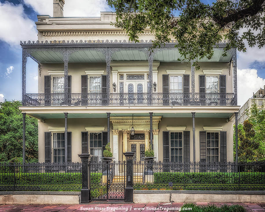 A two‑story historic building with a light exterior features tall shuttered windows, decorative columns framing the entrance, and ornate wrought‑iron balconies and railings. A black iron fence encloses the neatly kept front garden, creating a striking contrast between the light façade and the dark ironwork.