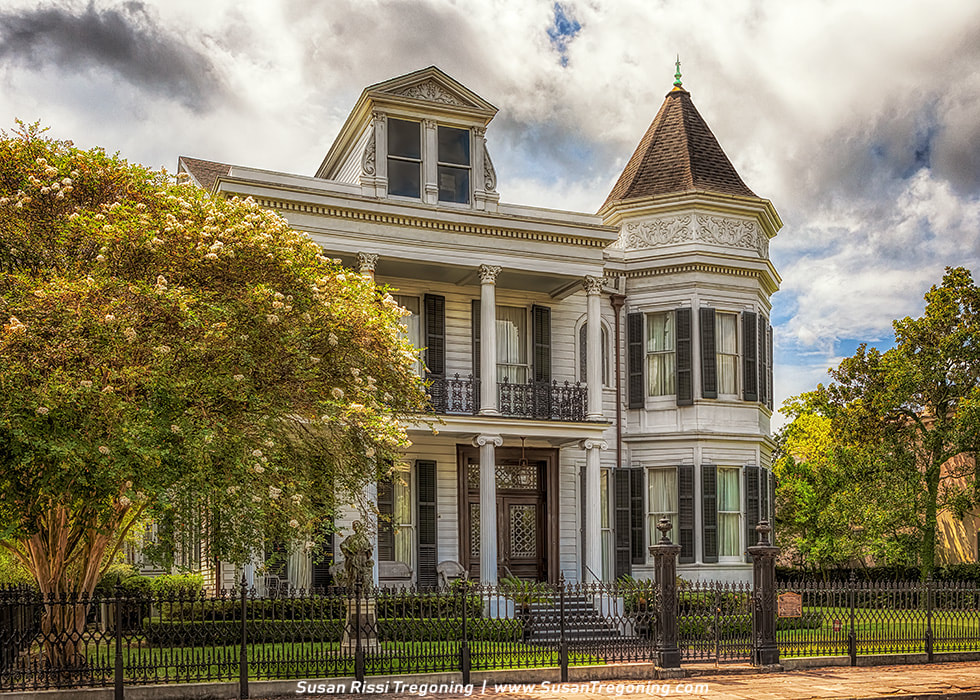 A large white Victorian‑style house features tall columns, an upper balcony, and a distinctive turret with a steep roof. Decorative molding and dark shutters highlight the façade, while a wrought‑iron fence and lush greenery—including a flowering tree—frame the property beneath a partly cloudy sky.
