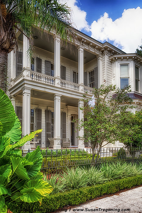 A large two‑story house with tall white columns and a second‑story balcony features shuttered windows, detailed roofline moldings, and light neutral exterior tones. A well‑maintained garden with tropical plants, trimmed hedges, and a wrought‑iron fence surrounds the home, with a partly cloudy sky overhead.