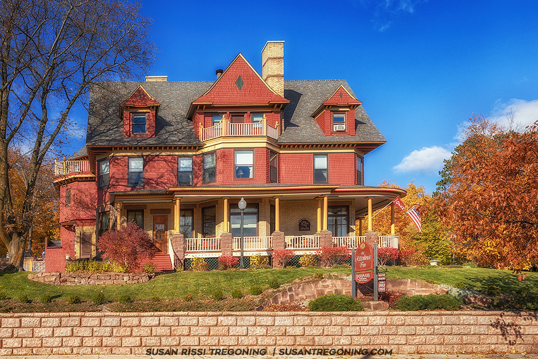 A large Victorian house with red and tan exterior walls, steep gabled roofs, and multiple dormers stands behind a stone retaining wall. A wraparound porch with yellow columns and decorative railings extends across the front. A sign in the yard reads “Redstone Inn” with “Luxury Inn” below it. An American flag hangs from the porch, and autumn trees with orange and yellow leaves surround the house under a bright blue sky.