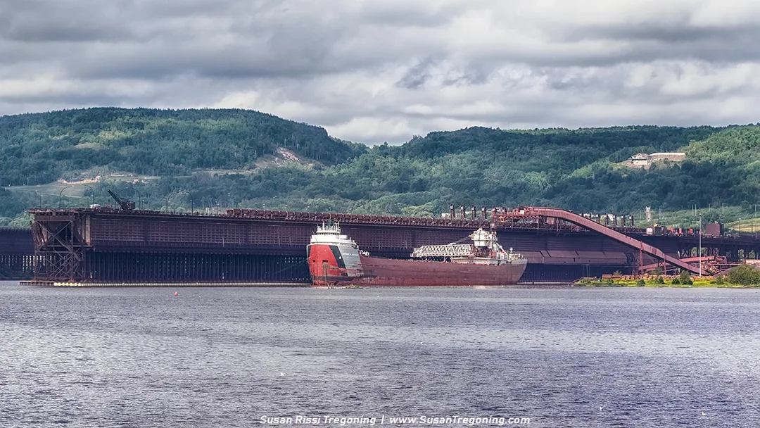 A large Great Lakes freighter, the Arthur M. Anderson, sits alongside an ore dock in long‑term layup. The ship’s hull stretches across the frame, with industrial structures and dock equipment visible behind it under bright, partly cloudy light.