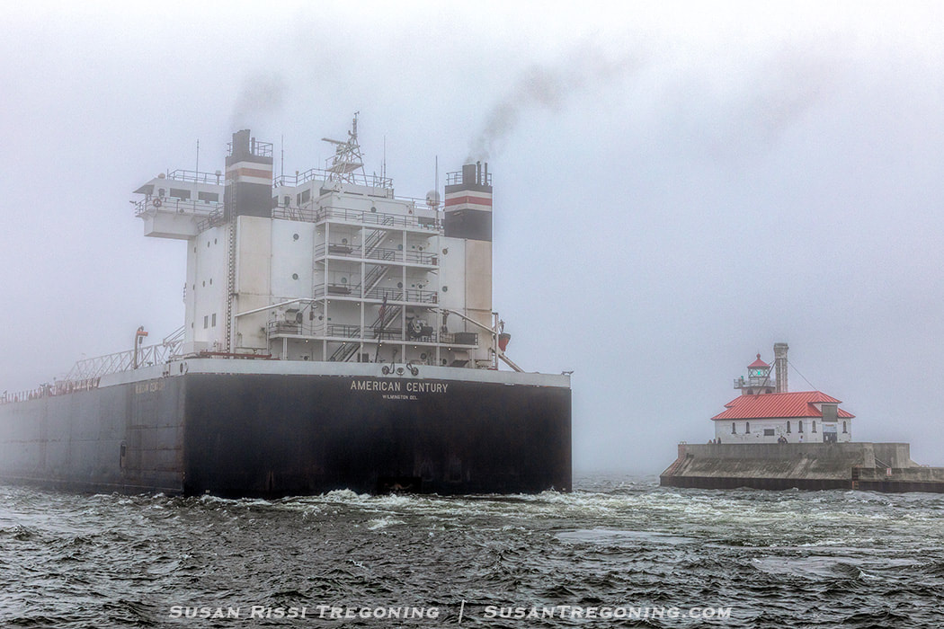 A large cargo ship, the American Century, moves through fog on Lake Superior as it passes the South Breakwater Outer Light at the end of the Duluth Ship Canal. The ship emits dark exhaust, and the lighthouse stands on a concrete pier to the right. The water is choppy under the gray, misty sky.