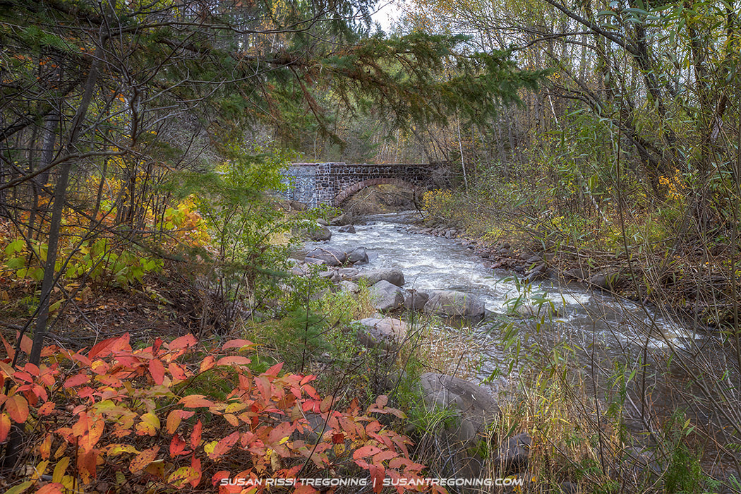 A small stone arch bridge crosses a rocky creek surrounded by dense autumn foliage in shades of red, orange, yellow, and green. Water flows over the rocks beneath the bridge, framed by trees and shrubs in a forest setting.