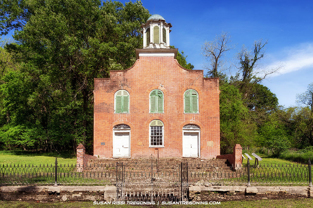 A historic brick building with a curved gable, two arched doorways, and three arched windows with green shutters stands behind a wrought‑iron fence, surrounded by lush green trees and grass in bright daylight.
