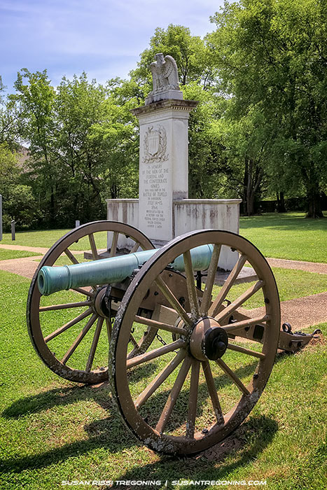 A historic cannon with wooden wheels stands in front of a tall stone monument topped with an eagle at Tupelo National Battlefield. The monument rises from a grassy area surrounded by trees, with a paved walkway leading toward it. Sunlight filters through the foliage, illuminating the cannon and memorial.