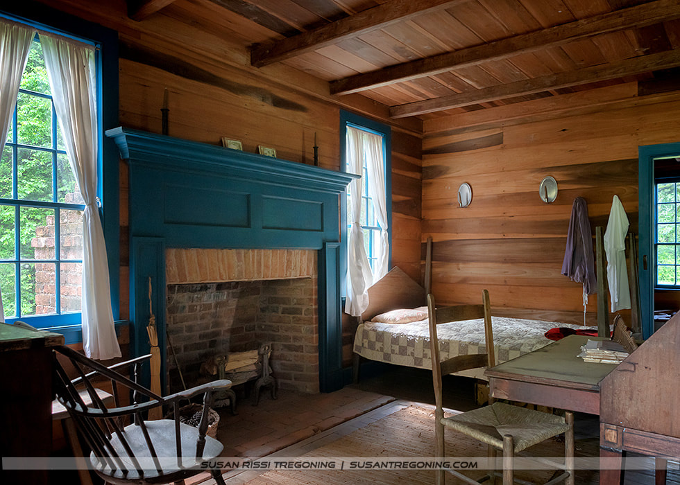A rustic 18th‑century bedroom features wood‑plank walls, exposed ceiling beams, and a brick fireplace with a blue-painted mantel. A simple bed with a patchwork quilt sits near large windows trimmed in blue, with white curtains filtering natural light. A wooden desk with papers, a chair, and a rocking chair stand near the hearth, while clothing hangs from bedposts and wall pegs, reflecting everyday life in the historic home.