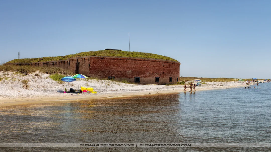 A historic brick fort sits along a sandy beach with people walking, swimming, and sitting under colorful umbrellas near the shoreline. The fort’s red brick walls rise above the sand, topped with grass, beneath a clear blue sky.