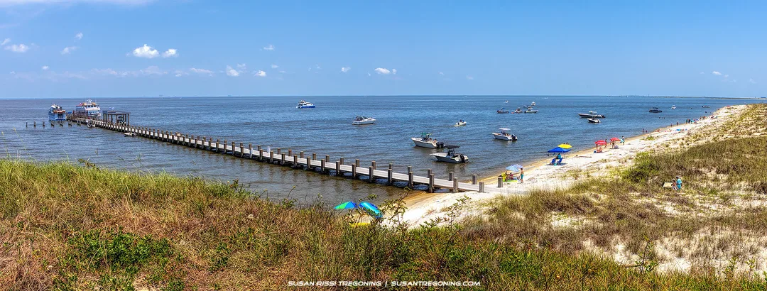 A long wooden pier extends over calm blue water with several anchored boats nearby. People sit under colorful umbrellas and relax along the sandy beach, with grassy dunes in the foreground and a clear sky above.
