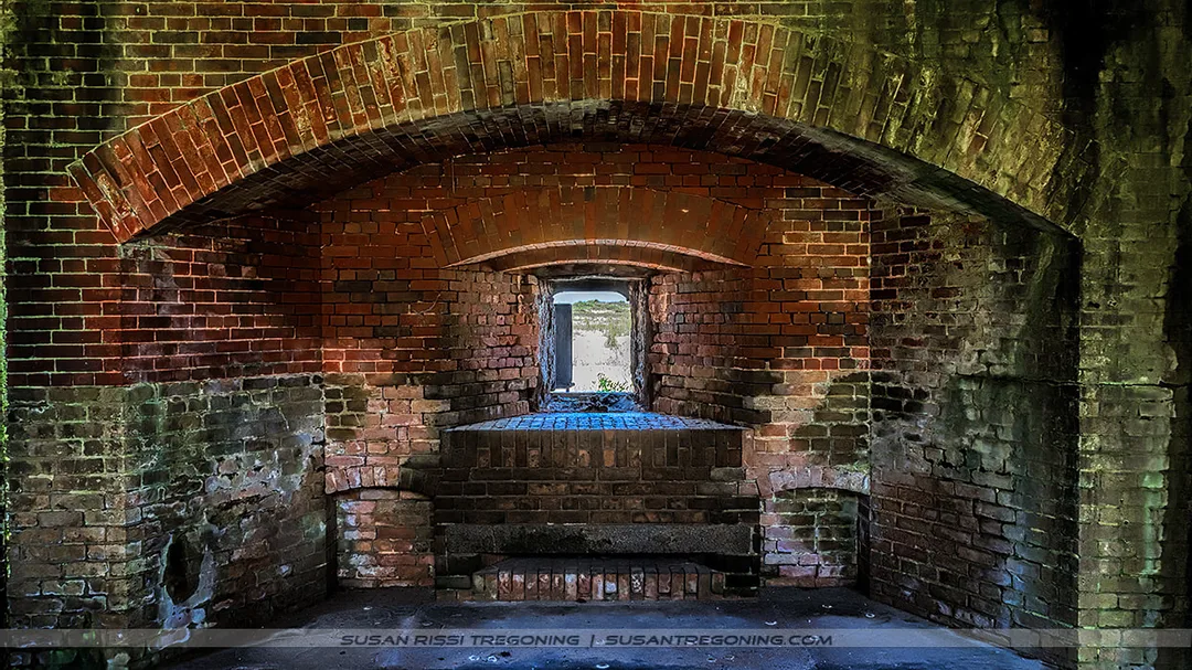 A brick casemate inside a historic fort features a series of arched ceilings and walls leading toward a bright rectangular opening at the far end. Natural light highlights the weathered brick and the shadowed interior passage.