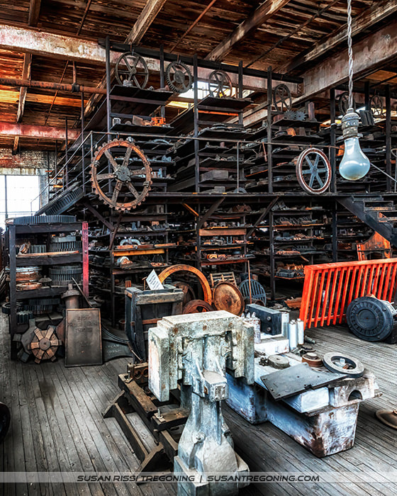 Shelves inside a historic workshop filled with metal patterns and mechanical parts, including gears, pulleys, and wheels stacked in rows. The space features worn wooden floors, aged brick walls, and a single hanging light bulb illuminating the textures of the metal and wood. The scene captures the dense, organized storage typical of an early industrial pattern shop.