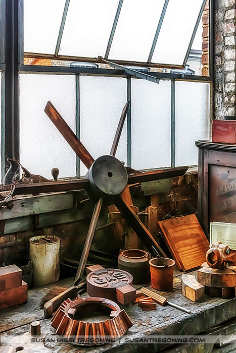 A six‑bladed wooden propeller leans against a frosted‑glass window inside a historic workshop. Smaller wooden molds, cylindrical forms, and geometric casting pieces are arranged on the worn workbench around it. The brick wall, aged surfaces, and scattered tools create an early manufacturing or pattern‑shop atmosphere.