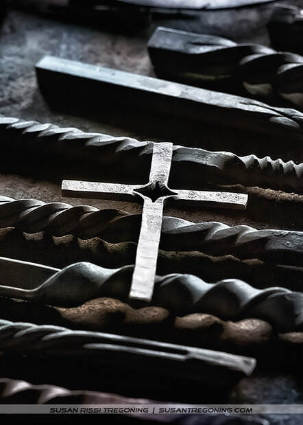 A collection of forged metal pieces arranged on a surface, including a central metal cross with a star‑shaped cutout and several twisted or textured metal rods placed around it. The lighting highlights the metallic surfaces and craftsmanship, giving the scene a handcrafted, blacksmith‑workshop feel.