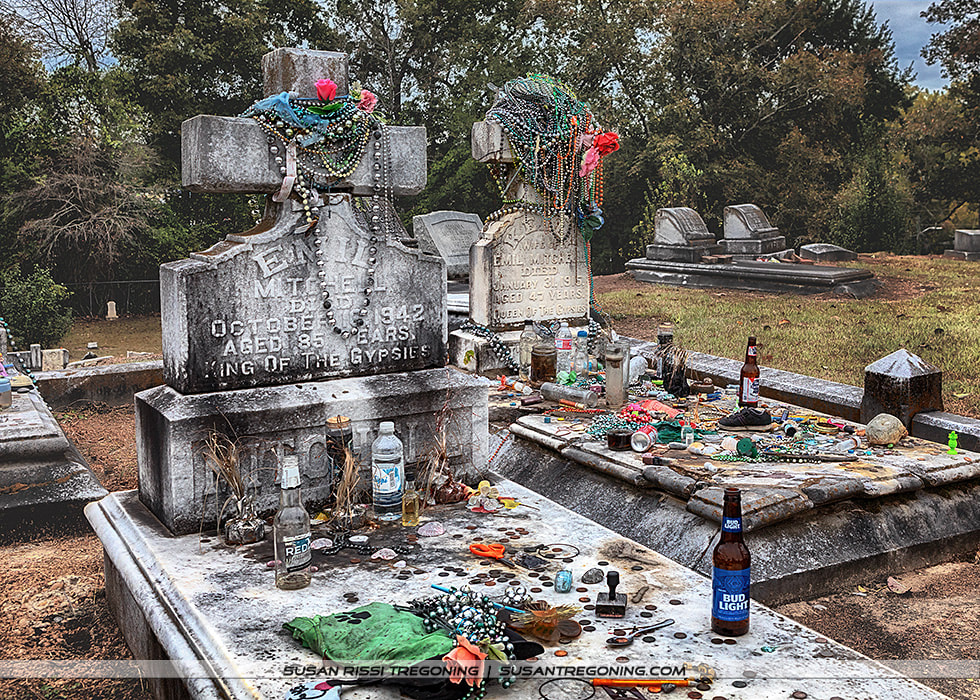 Two ornate stone cross‑shaped headstones stand side by side in a cemetery, each covered with colorful beads, necklaces, coins, bottles, and other offerings. The left marker is inscribed for Emil Mitchell, and the right for Emilia Mitchell. Grass, trees, and additional gravestones appear in the background, creating a quiet, weathered memorial scene.