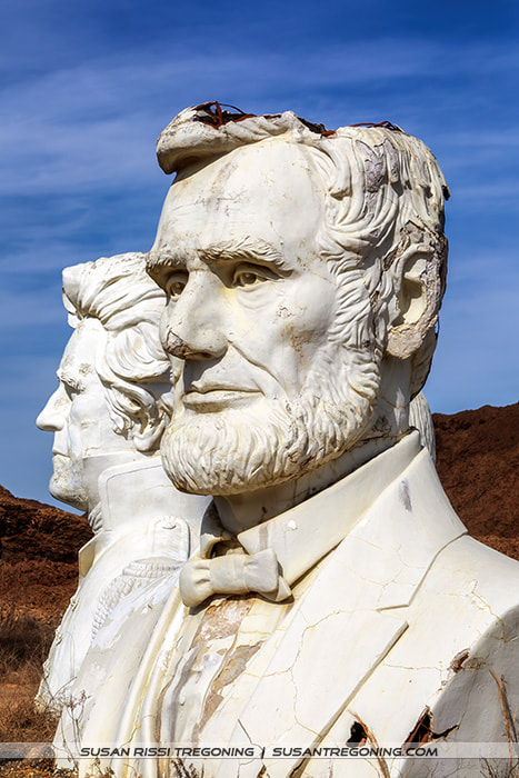 A large, weathered concrete bust of Abraham Lincoln stands in the foreground, with another presidential head positioned behind it. Both sculptures show visible cracks and discoloration, set outdoors against rocky terrain and a clear blue sky.