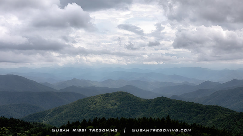 Layered blue mountain ridges stretch into the distance at Cowee Mountain Overlook as storm clouds move in. Dense green forest fills the foreground, and the fading ridgelines recede into a hazy, overcast sky.