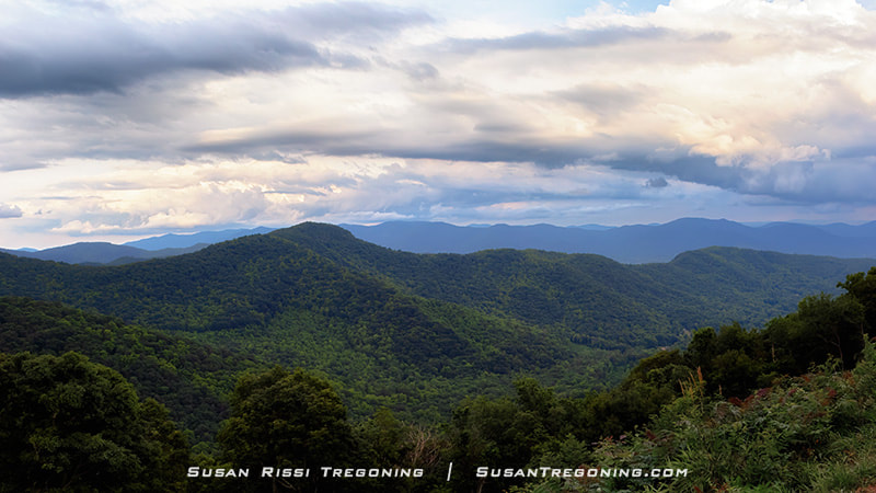 A wide mountain panorama is seen from Lane Pinnacle Overlook, with layers of blue ridges fading into the distance under early‑evening light.