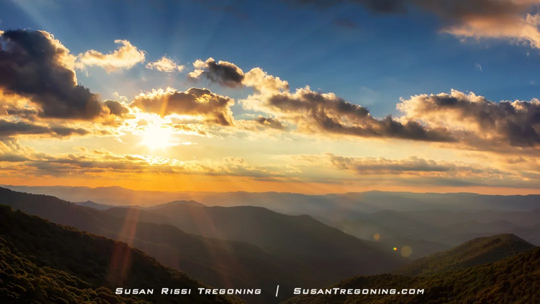 The sun sits low in the sky above the Blue Ridge Mountains near Craggy Gardens, with bright golden light radiating outward in multiple directions. Sunbeams stream through breaks in the clouds, creating a dramatic ring of rays across the sky. Layered mountain ridges fade into the distance, shifting from warm tones near the horizon to cooler blue hues farther away.