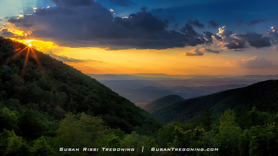 The sun sets behind a mountain ridge at Taylors Mountain Overlook, with warm rays shining through breaks in the clouds. Forested hills fill the foreground, and distant blue mountain layers fade toward the horizon beneath a partly cloudy sky.