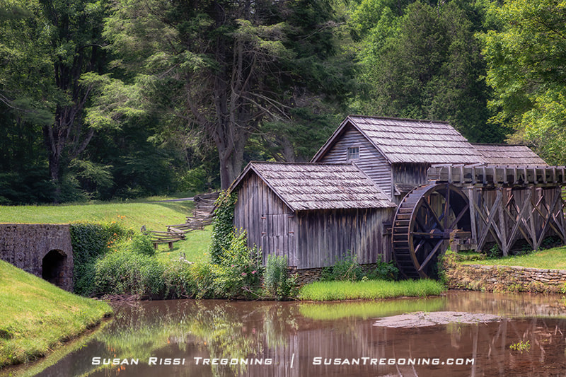 A rustic wooden watermill with a large water wheel sits beside a still pond, surrounded by lush green trees. The weathered building reflects in the water, and a stone culvert is visible to the left where water flows beneath it.