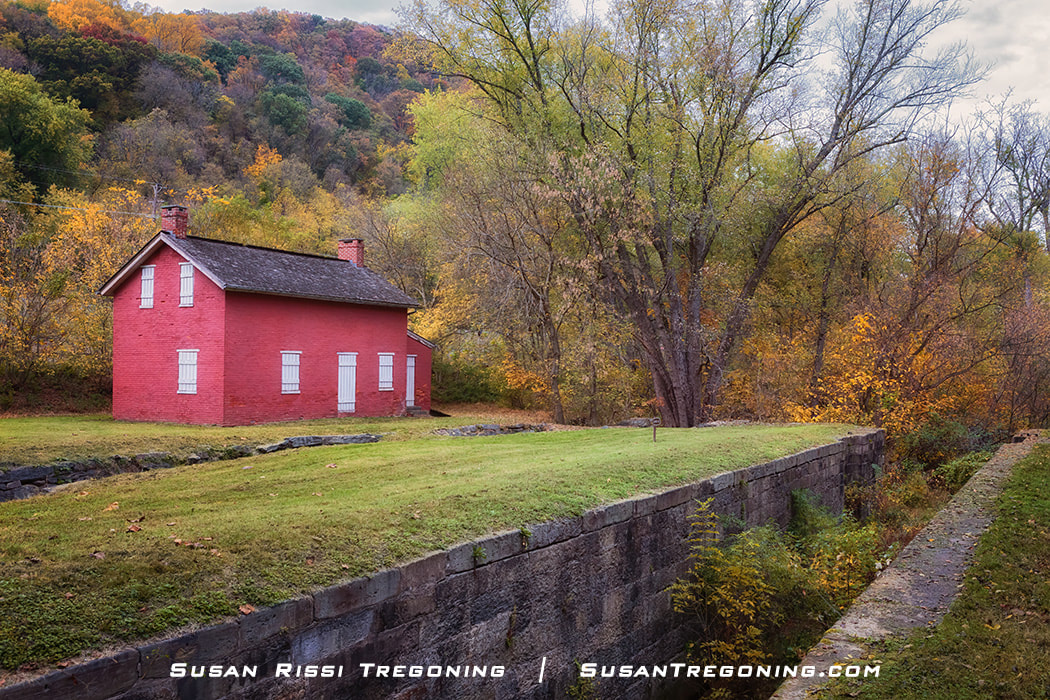 A small red‑brick lockhouse with white shutters stands beside a grassy canal bed and a low stone wall, surrounded by autumn trees in shades of green, yellow, and orange. Forested hills rise in the background, creating a peaceful fall landscape.