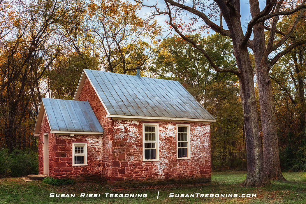A small historic brick schoolhouse with a weathered whitewashed exterior and a gray metal roof stands among tall autumn trees. The building has white‑trimmed windows and a door on the left, surrounded by warm fall foliage.