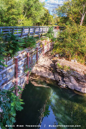 A historic stone aqueduct spans a calm, tree‑lined waterway, its metal supports and deck partially shaded by dense green foliage, with sunlight reflecting off the water below.