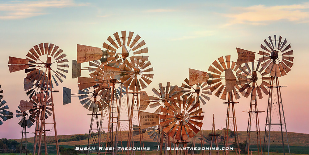Several vintage metal windmills stand in a grassy field at sunset, each with weathered blades and tail vanes. Rolling hills and a colorful sky fill the background.