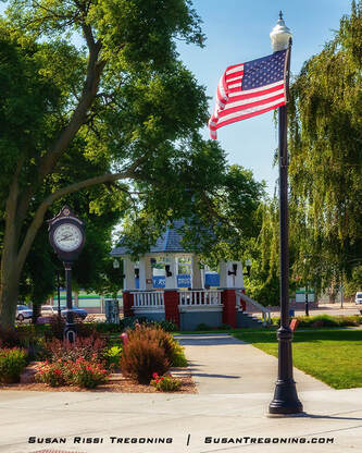 A park scene shows a lamppost with an American flag, a decorative street clock, and a gazebo surrounded by trees and landscaped flower beds. Paved walkways and green lawn areas fill the town square.
