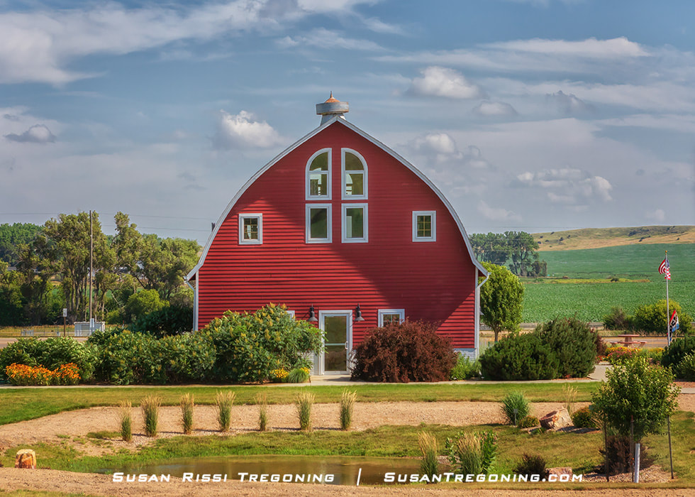 A bright red barn with white trim stands in a landscaped area with shrubs, small trees, and a gravel path. Green fields and rolling hills stretch into the distance beneath a partly cloudy blue sky.