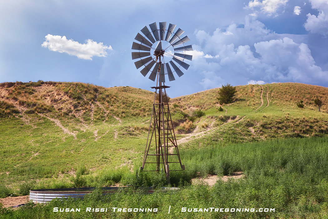 A tall metal windmill stands in a grassy area beside a round metal stock tank, with rolling hills and scattered shrubs in the background under a partly cloudy sky.
