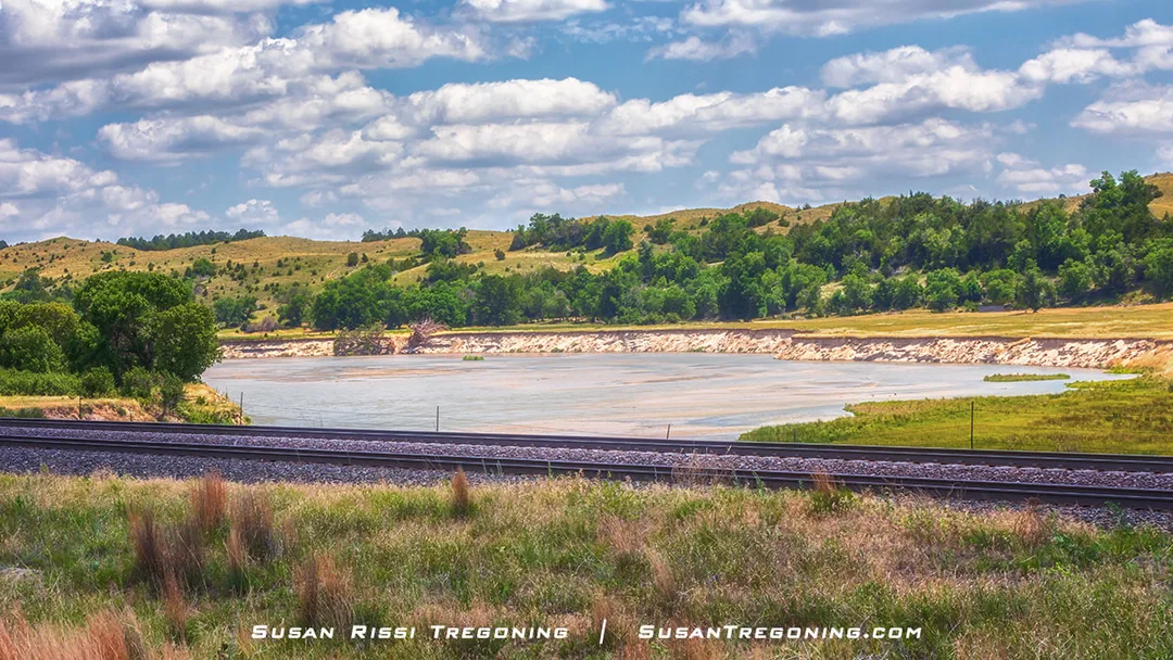 A river curves through green vegetation with rolling grass‑covered hills and scattered trees in the distance under a partly cloudy sky.