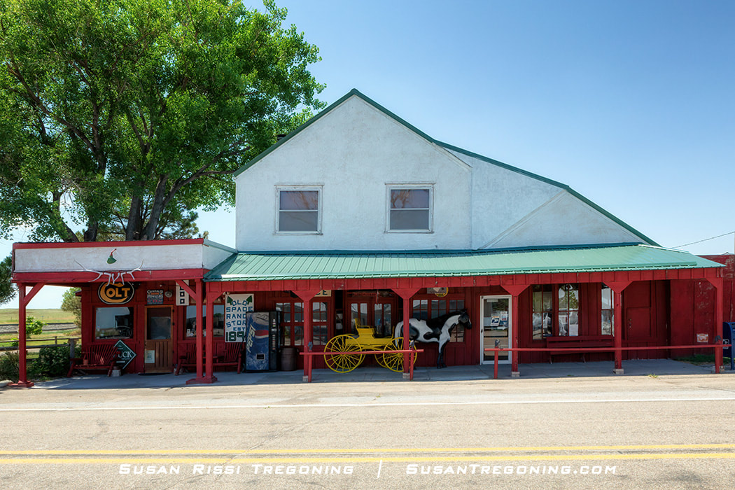 A rustic building with a white upper facade and red lower walls sits along a paved road, with signs reading “Old Spade Ranch Store 1891.” A yellow horse‑drawn carriage and a black‑and‑white horse statue are displayed on the porch, with large trees providing shade on one side.

