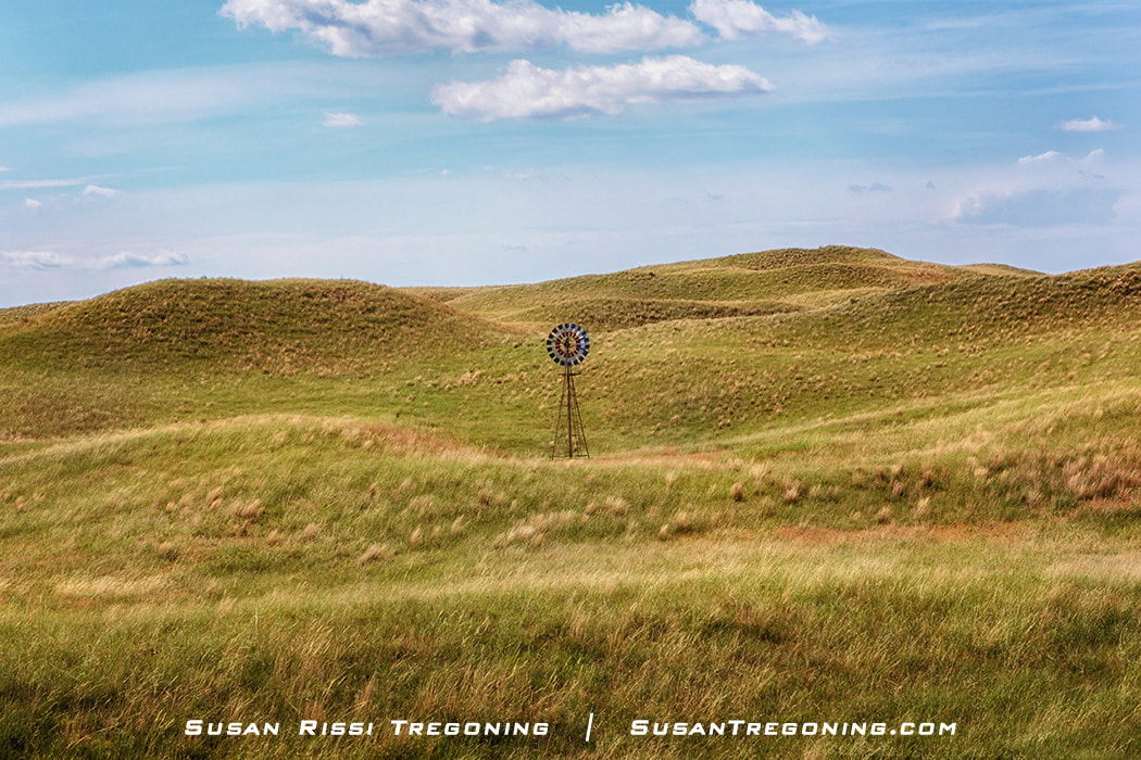 A windmill painted in red, white, and blue stands on a grassy hill surrounded by rolling golden‑green hills under a partly cloudy blue sky.