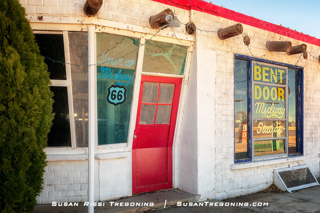 The Bent Door Café in Adrian, Texas, with a slanted red door on a white brick building, large windows displaying colorful Midway Station signage, red trim along the roofline, and string lights hanging across the front.