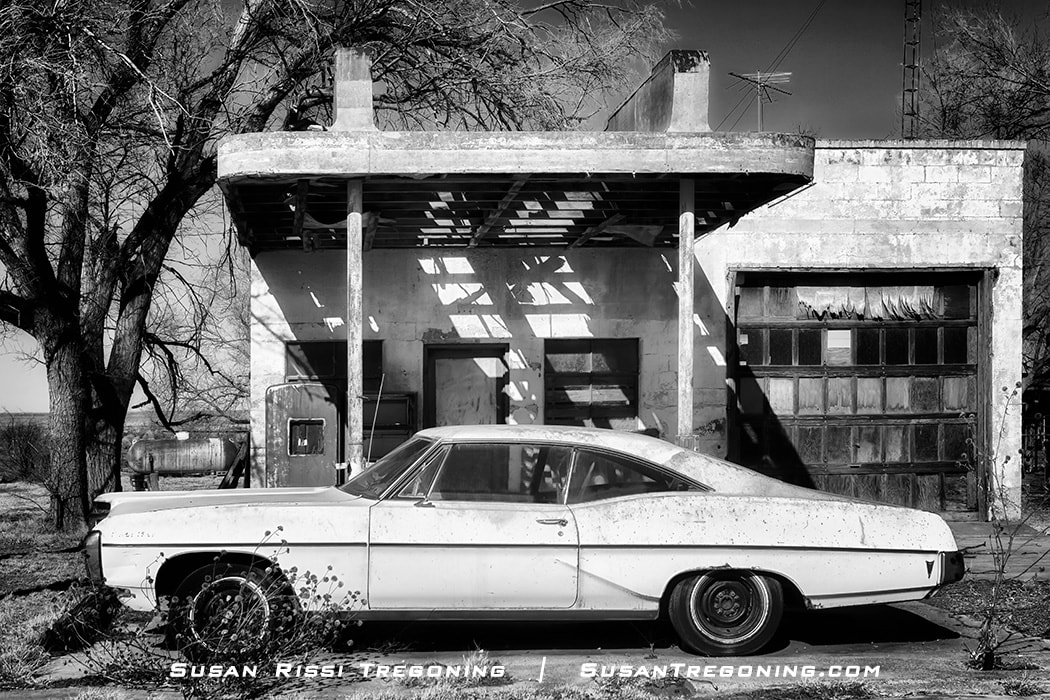 A weathered 1968 Pontiac Catalina parked in front of a dilapidated Texaco Service Station in Glenrio, Texas, with a partially collapsed roof supported by two columns, broken garage‑door windows, and leafless trees surrounding the abandoned structure.
