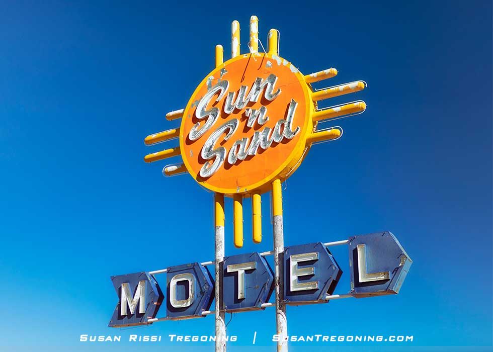 The Sun ’n Sand Motel sign in Santa Rosa, New Mexico, featuring a bright orange circular top with yellow rays in a Zia‑influenced design and white script lettering. Blue geometric panels below spell out “Motel,” all set against a clear blue sky.