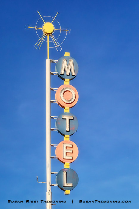 A tall vintage motel sign in Albuquerque with circular blue and pink panels stacked vertically to spell “MOTEL,” topped by a yellow sunburst‑shaped ornament, standing against a clear blue sky.