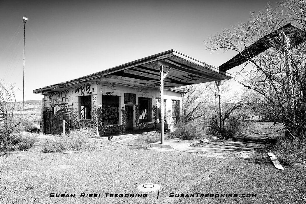 An abandoned gas station structure with a large overhanging roof, broken windows, and graffiti‑covered walls stands in a dry landscape. Sparse shrubs surround the building, with a tall communication tower and distant hills visible under a clear sky.