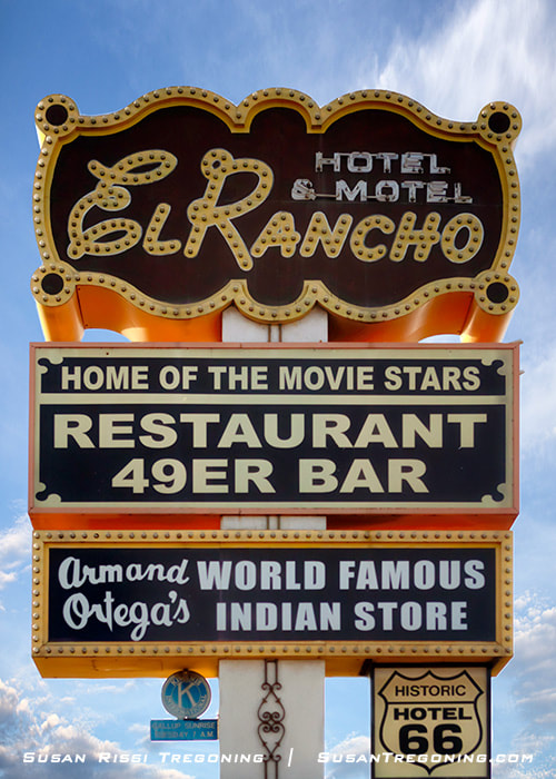 The historic El Rancho Hotel neon sign stands on a tall metal frame, with illuminated lettering spelling “El Rancho Hotel.” The sign has a classic vintage design and is set against the sky.