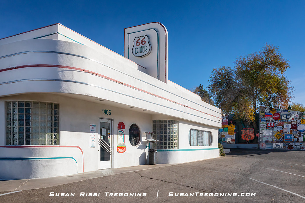 A white Art Deco‑style diner with rounded corners, horizontal colored stripes, and glass block windows. A vertical sign reads “66 Diner,” and the entrance displays the number 1405. A Coca‑Cola logo appears near the door, and a wall covered in vintage road signs is visible in the background under a clear blue sky.
