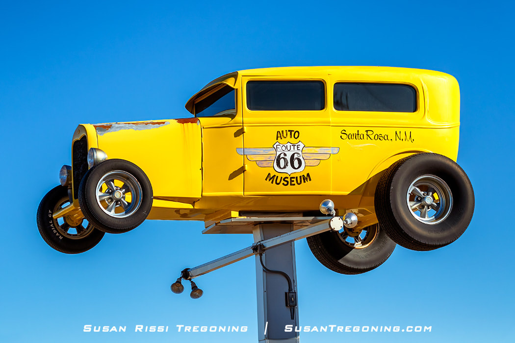 A bright yellow roadster mounted high on a post serves as signage for the Route 66 Auto Museum in Santa Rosa, New Mexico. The car has chrome wheels and visible rust on the front fender, set against a clear blue sky.